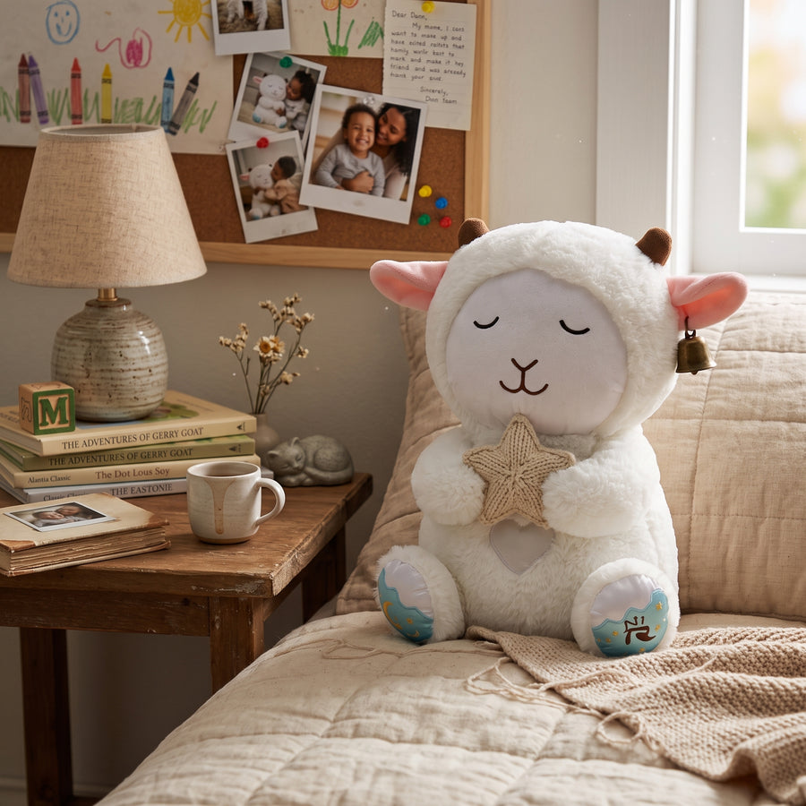 A plush sheep toy with a white fluffy body, pink inner ears, brown horns, embroidered closed eyes, a beige star-shaped nose, and small blue-and-white patterned feet sits on a beige quilted bed in a softly styled bedroom. A wooden bedside table with a lamp, mug, books, and small decor is beside it, and a cork board with photos and drawings is on the wall behind.