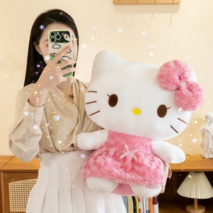 A woman taking a mirror selfie holds a large Hello Kitty plush toy wearing a pink fluffy dress and bow. The photo is taken indoors in a warm room with decorative sparkles and hearts overlaid digitally.
